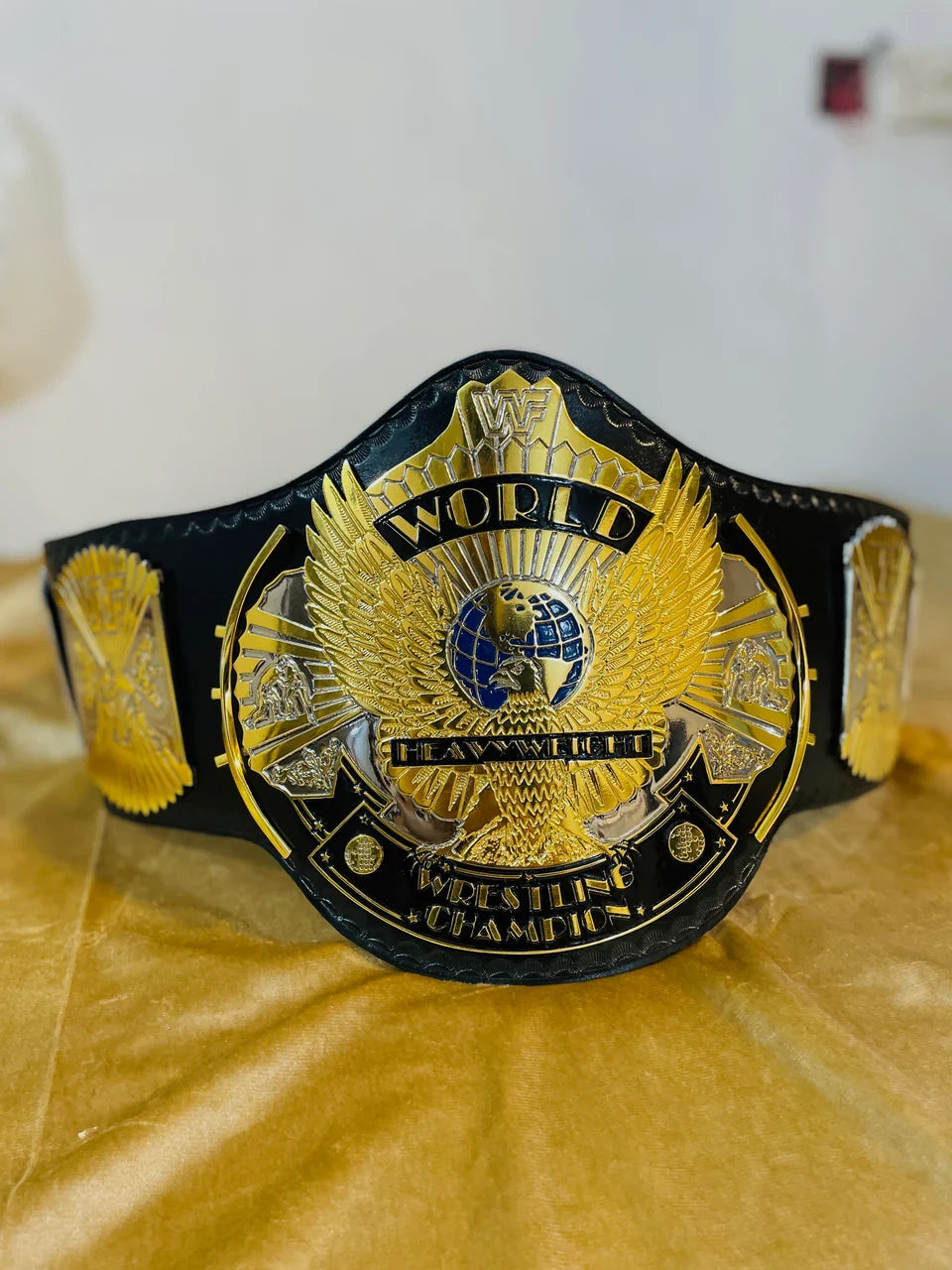 A black and gold championship wrestling belt with an eagle emblem and 'WORLD HEAVYWEIGHT BRAWLING CHAMPION' text, displayed on a gold fabric surface.